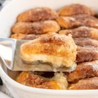 A dish of cinnamon sugar-coated dumplings in a white baking dish. One is being lifted out with a spatula. Green apples are visible in the background.