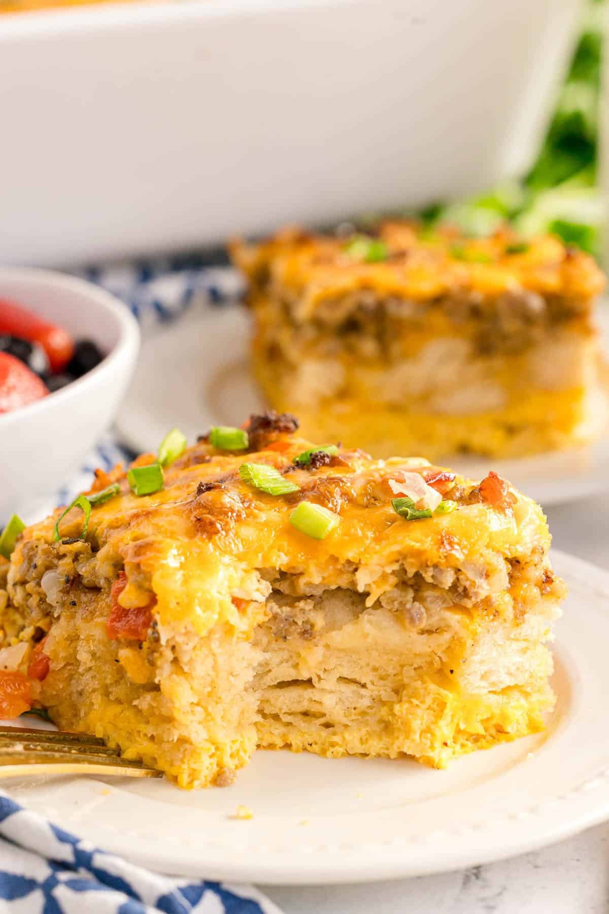 A slice of biscuit breakfast bake on a white plate with a bite missing, topped with melted cheese and green onions.
