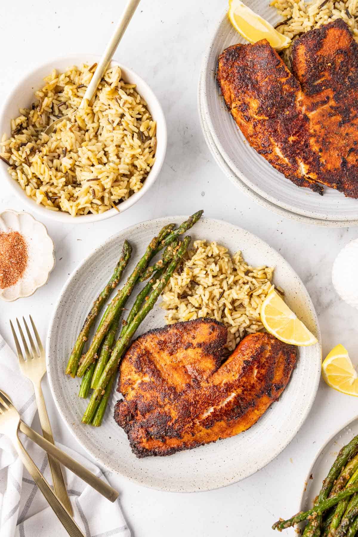 Overhead view of two plates with blackened tilapia, rice, and asparagus, with a bowl of rice on the side.