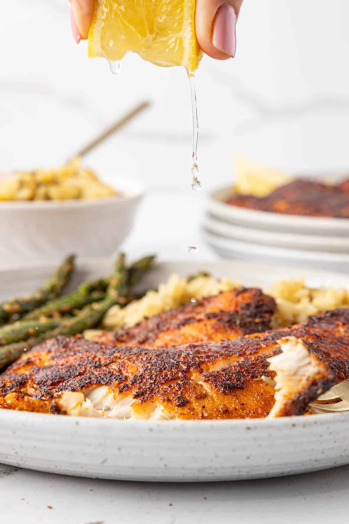 Lemon wedge being squeezed over a plate of blackened tilapia with asparagus and rice in the background.