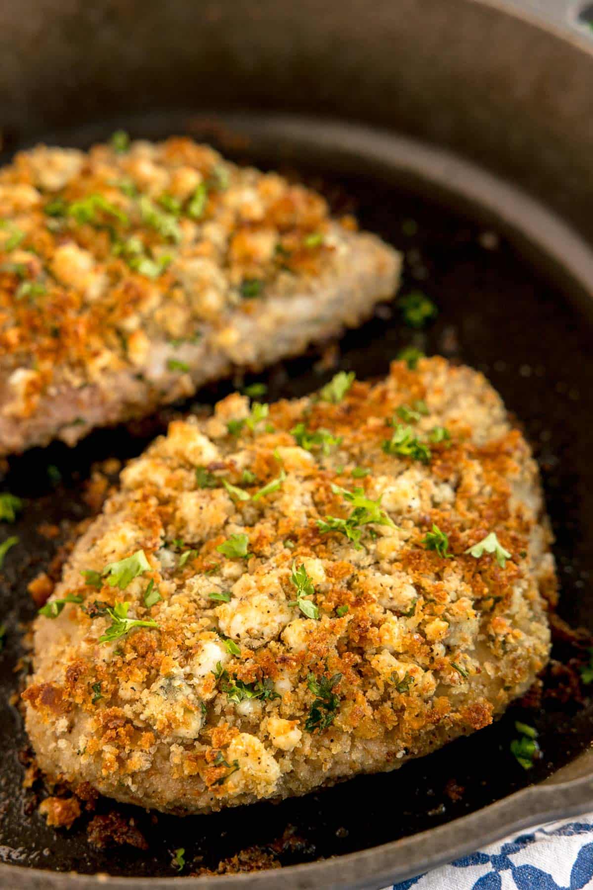 A close-up of a crusted pork chop in a cast iron skillet, topped with chopped parsley.