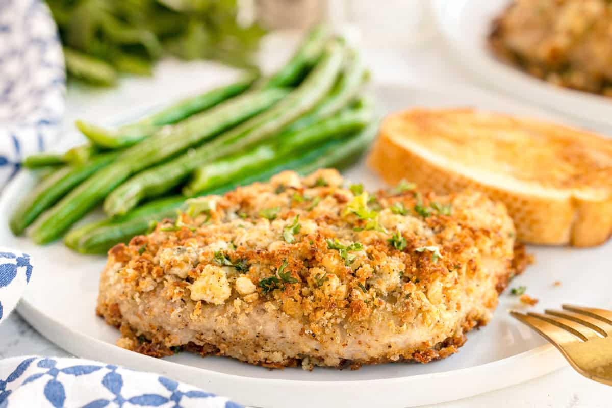 A plate with a blue cheese crusted pork chop, green beans, and toasted bread in the background.
