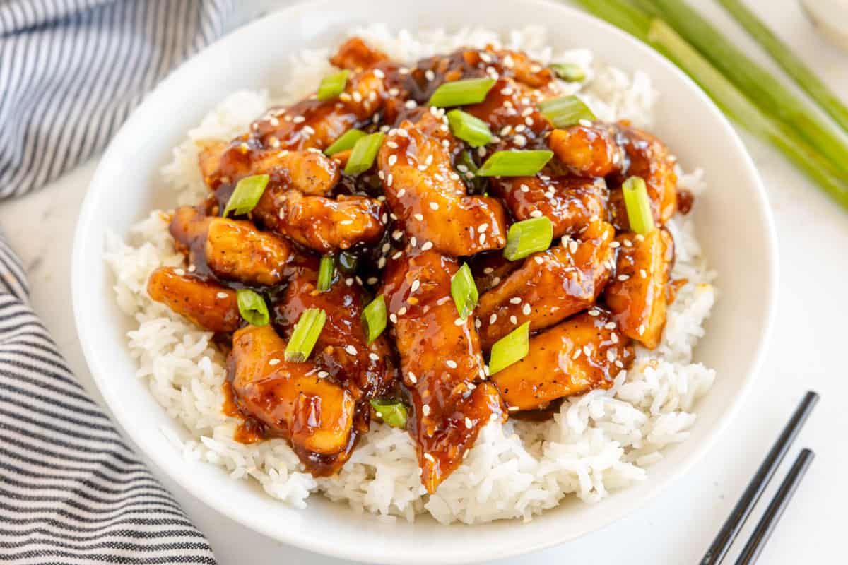 A bowl of white rice topped with pieces of cooked chicken in a dark sauce, garnished with sesame seeds and chopped green onions, placed next to a black and white striped cloth and chopsticks.