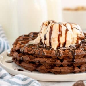 A stack of chocolate waffles topped with a scoop of vanilla ice cream and drizzled with chocolate syrup on a white plate.