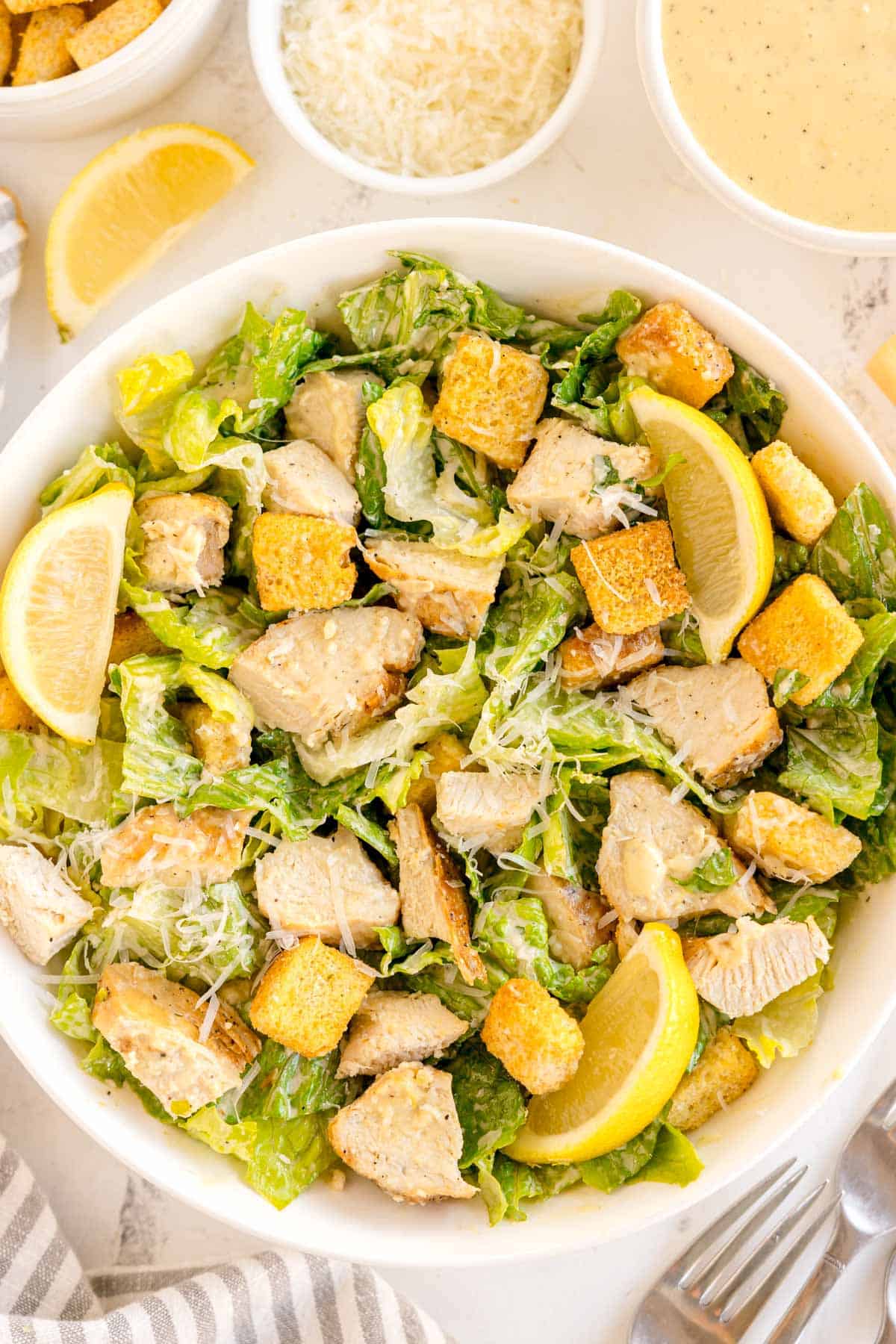 Chopped chicken Caesar salad with romaine, croutons, shredded cheese, and lemon wedges in a white bowl.