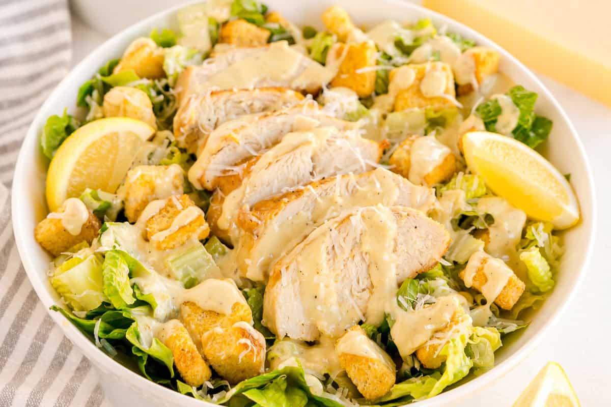 Caesar salad with sliced chicken, croutons, shredded cheese, lemon wedges, and creamy dressing.
