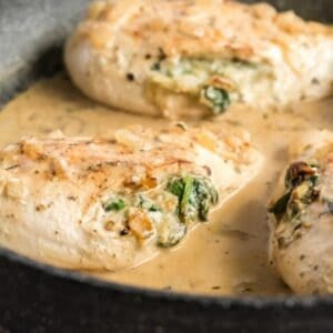 Two chicken breasts stuffed with spinach and cheese are cooking in a creamy sauce in a skillet.