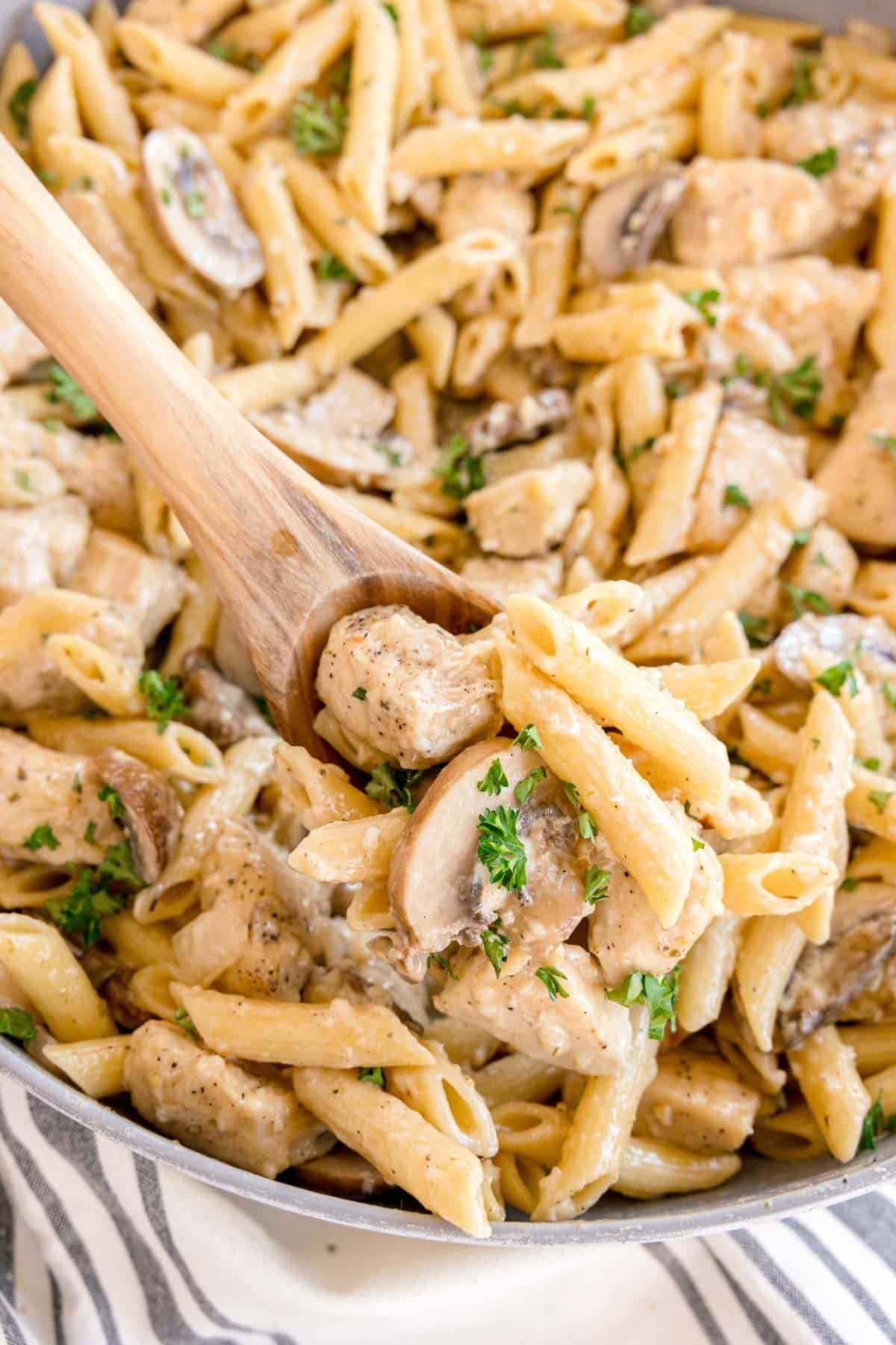 A wooden spoon lifting creamy penne pasta with chicken and mushrooms.