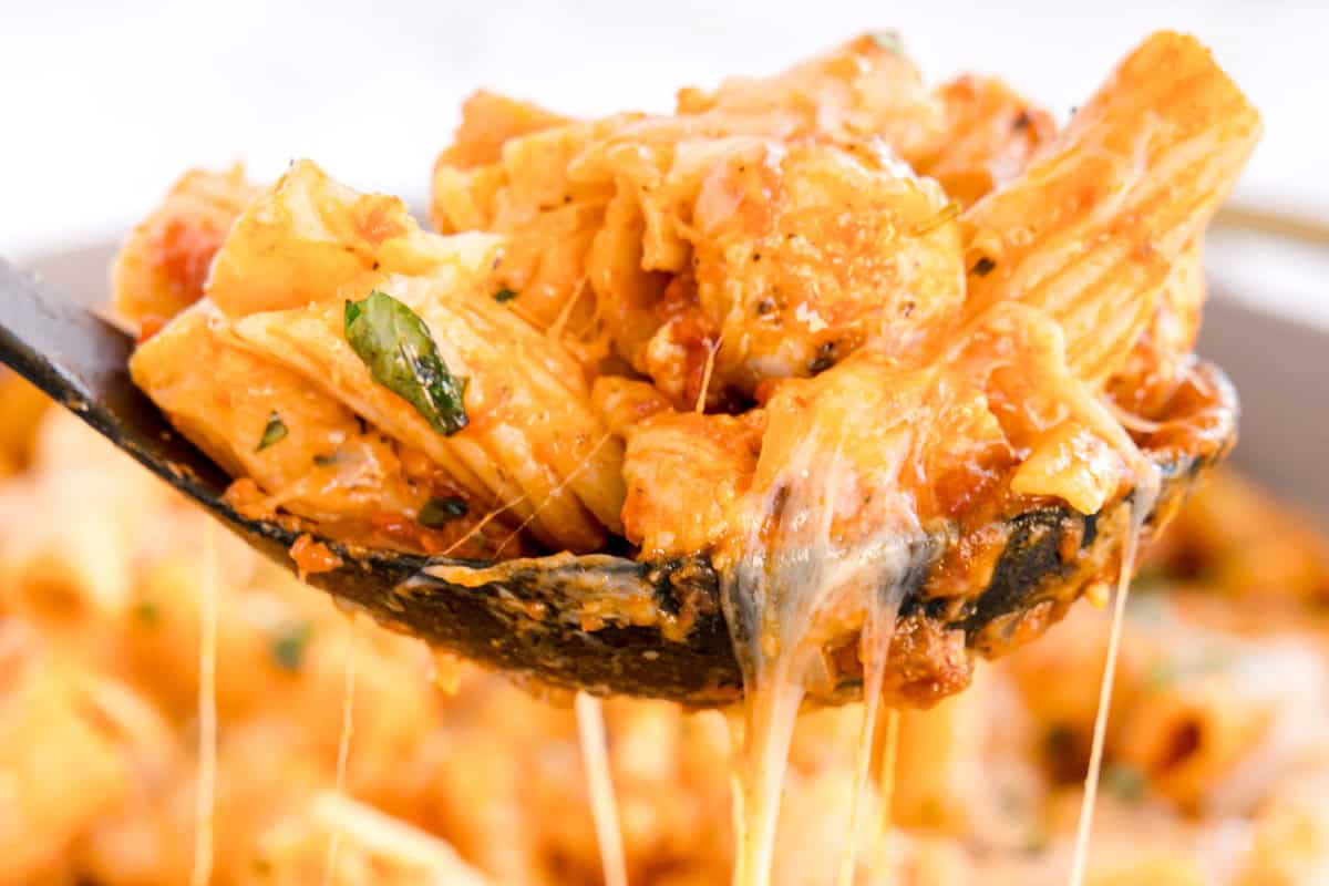 A ladle of rigatoni with chicken, marinara sauce, and melted cheese held over the pan.