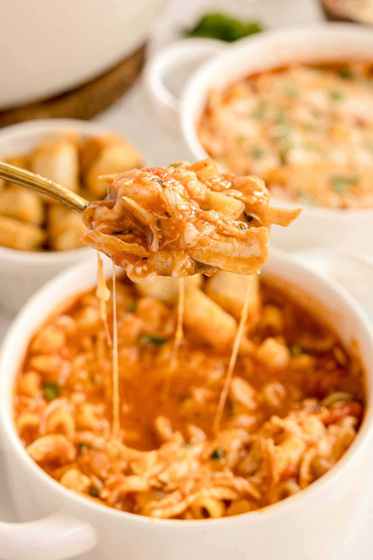 A spoonful of chicken parmesan soup with melted cheese lifted above a bowl filled with pasta and croutons.