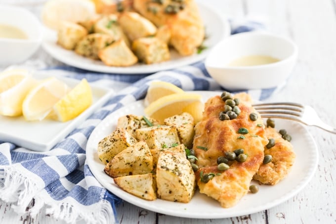 chicken piccata on a plate with roasted potatoes, and cut up lemon with a blue and white napkin