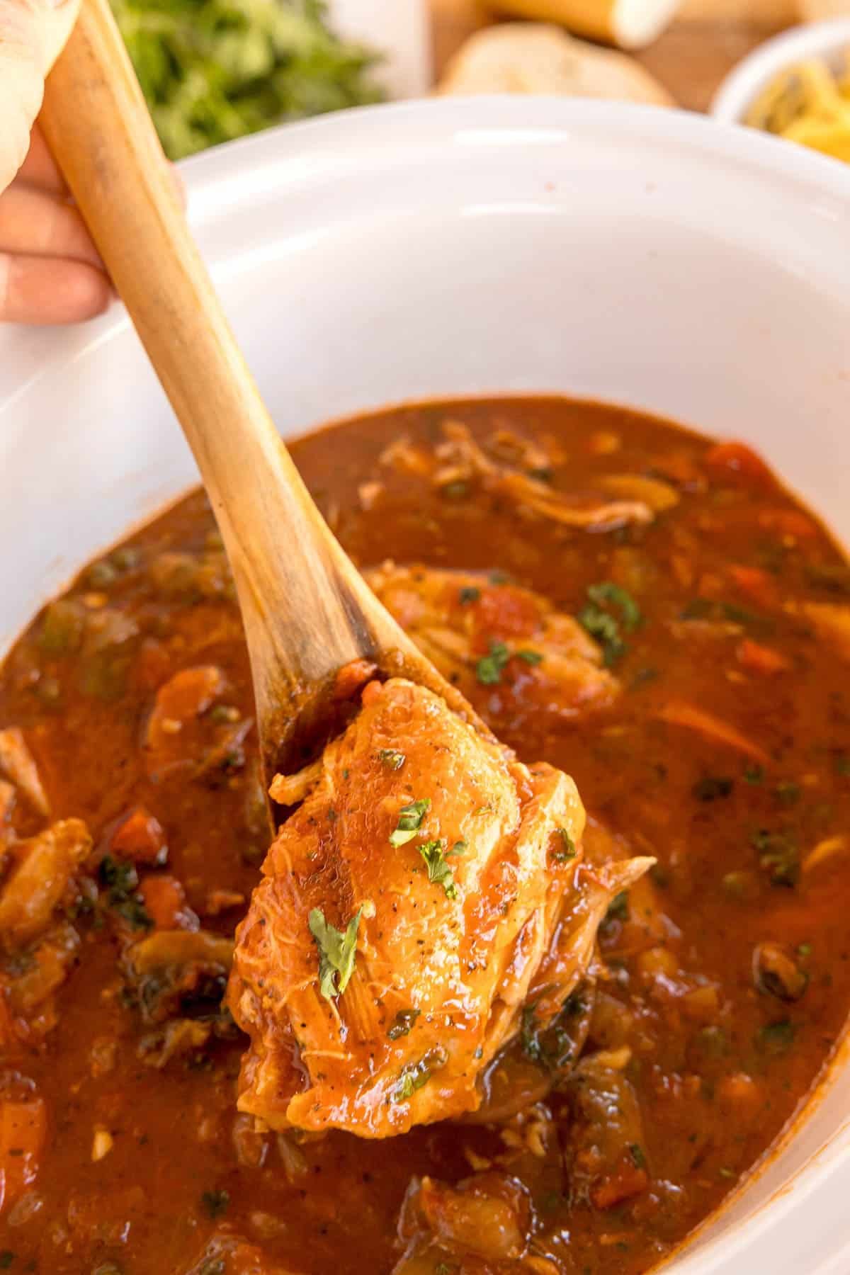A wooden spoon lifting a piece of tender chicken from a crockpot filled with rich, tomato-based chicken cacciatore sauce, garnished with fresh parsley.