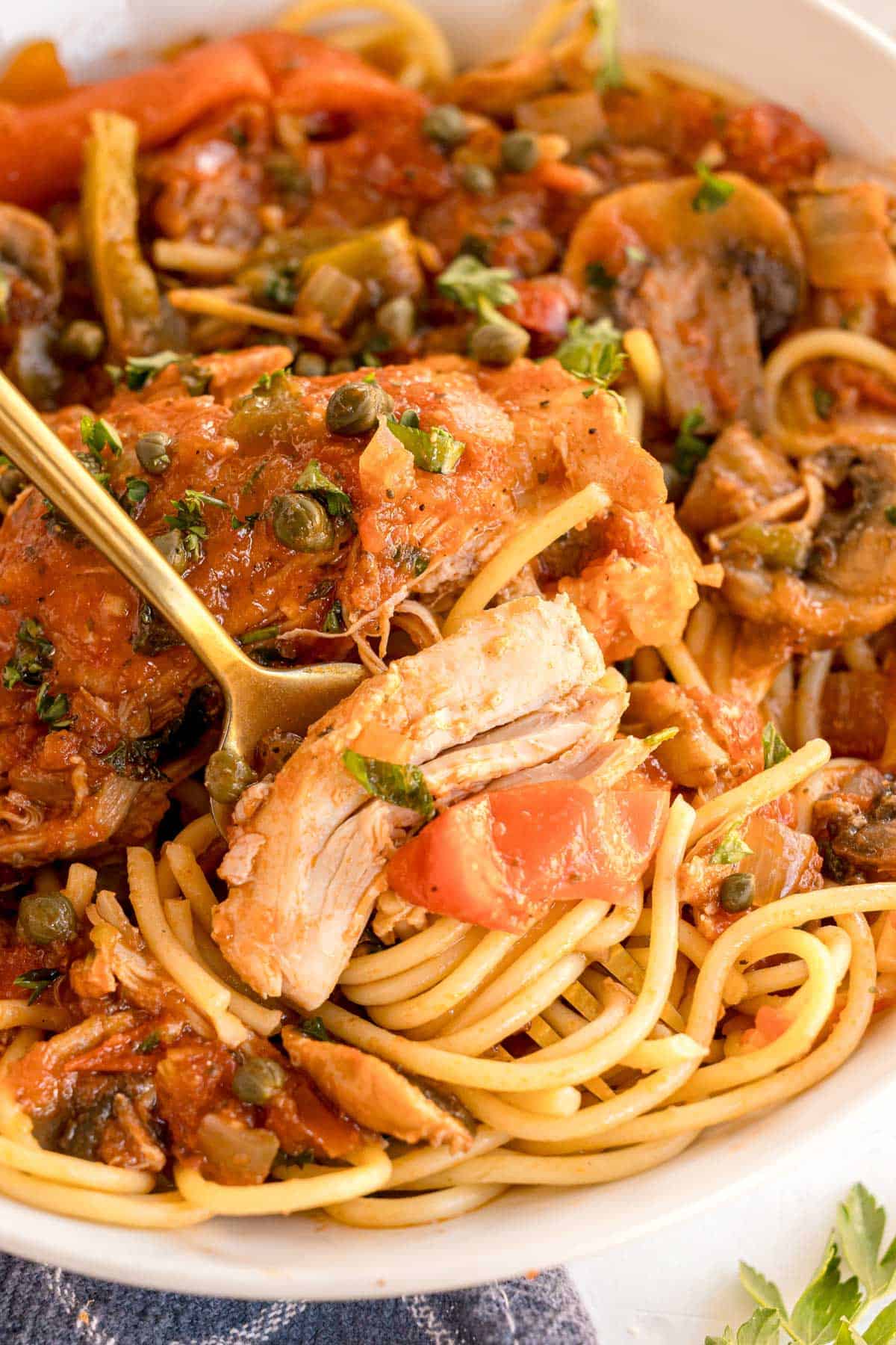 A close-up of a plate of spaghetti topped with chicken cacciatore, garnished with capers and fresh parsley, with a gold fork holding a piece of chicken.
