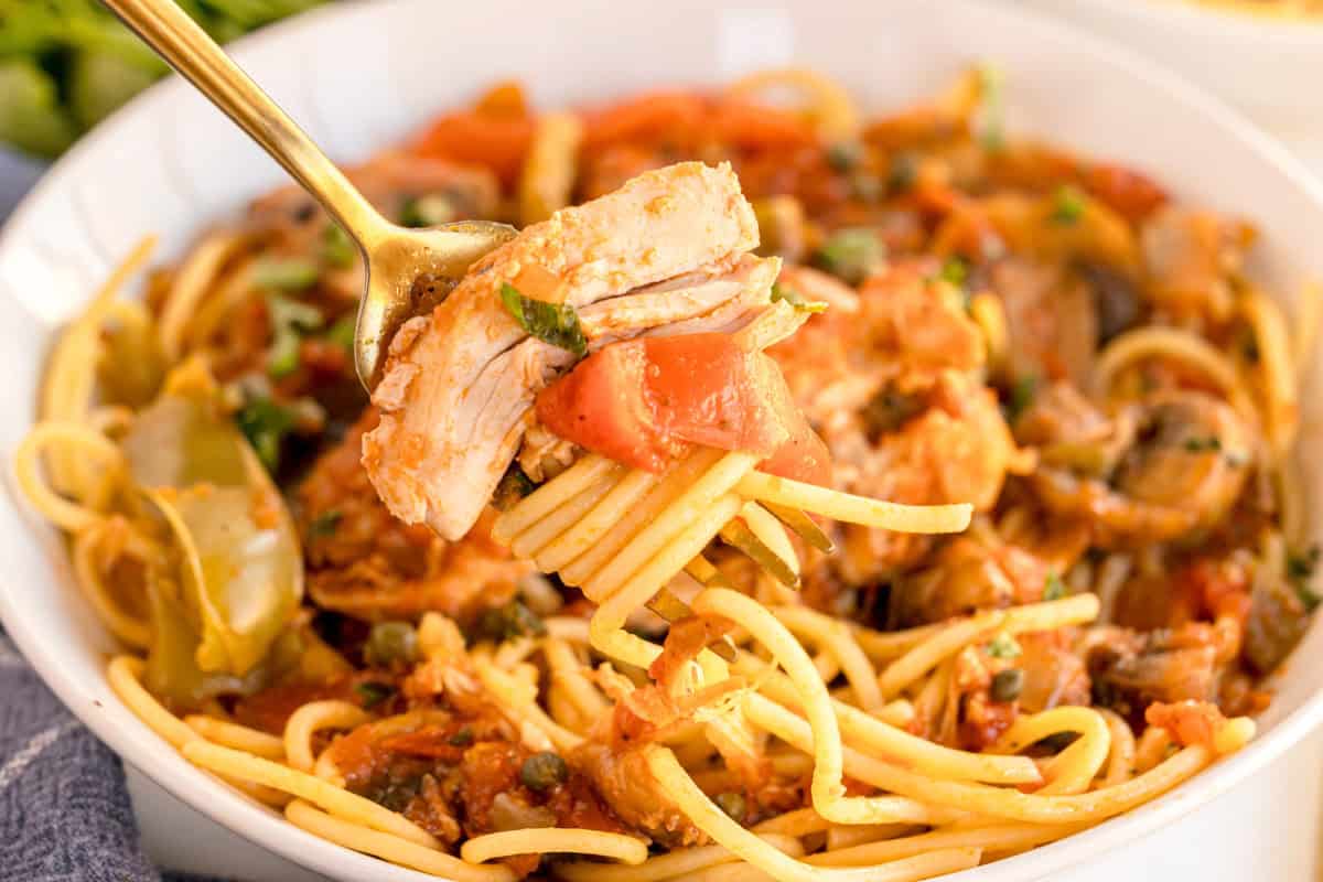 A fork lifting a piece of tender chicken with spaghetti, tomatoes, and capers, with a bowl of chicken cacciatore in the background.