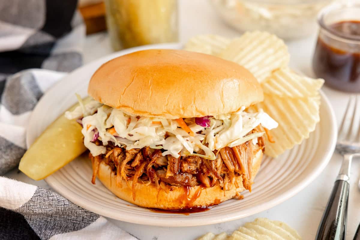A pulled pork sandwich topped with coleslaw sits on a white plate accompanied by potato chips and a pickle spear.