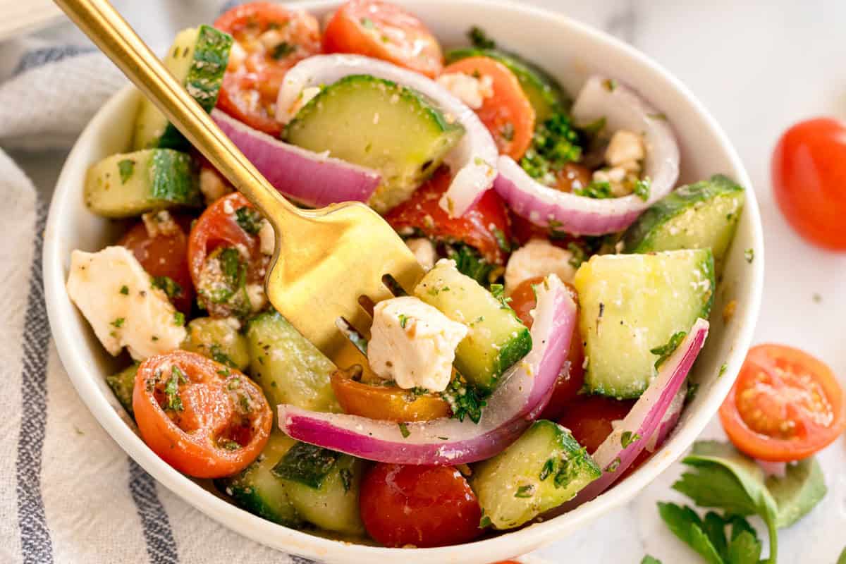 A fork holding a bite of cucumber salad with tomato, onion, and feta.