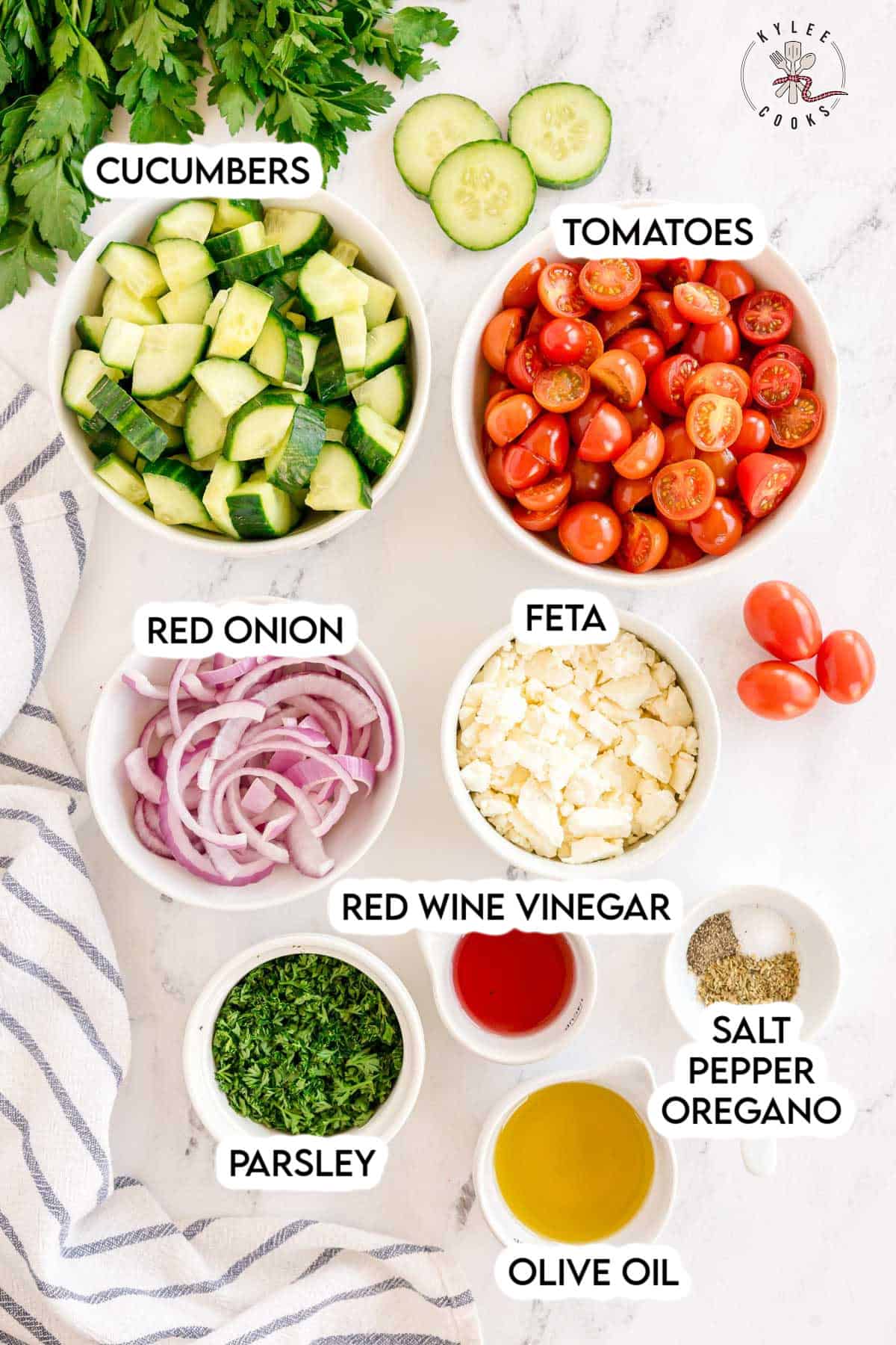 Bowls of cucumbers, tomatoes, red onion, feta, parsley, vinegar, oil, and seasonings. Text on the image says 'CUCUMBERS', 'TOMATOES', 'RED ONION', 'FETA', 'RED WINE VINEGAR', 'PARSLEY', 'OLIVE OIL', 'SALT PEPPER OREGANO'.