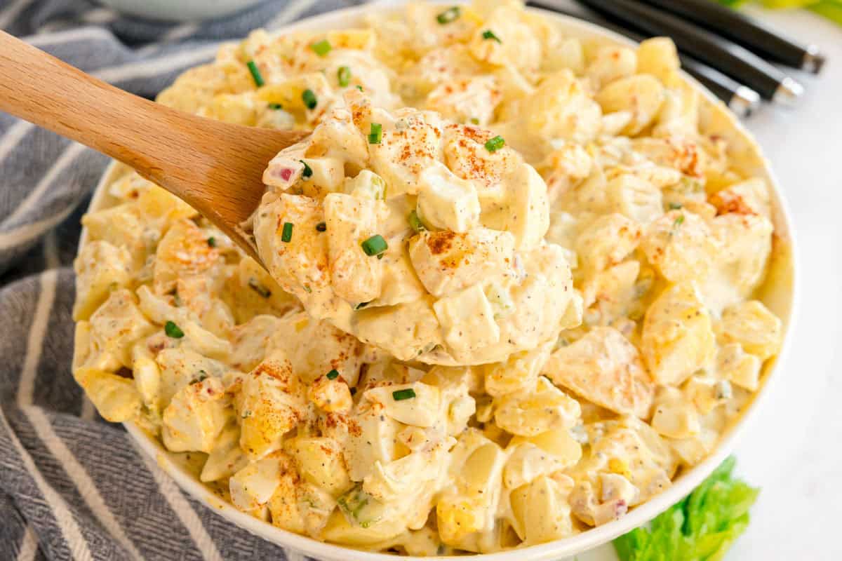 A wooden spoon lifting a scoop of creamy potato salad from a full bowl.