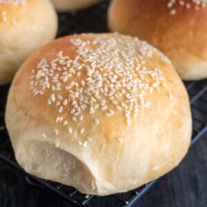 A close-up of a golden brown hamburger bun topped with sesame seeds, resting on a cooling rack with other buns in the background.