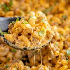 A close-up of a spoon scooping cheesy macaroni with ground meat and herbs, with melted cheese stretching from the spoon.