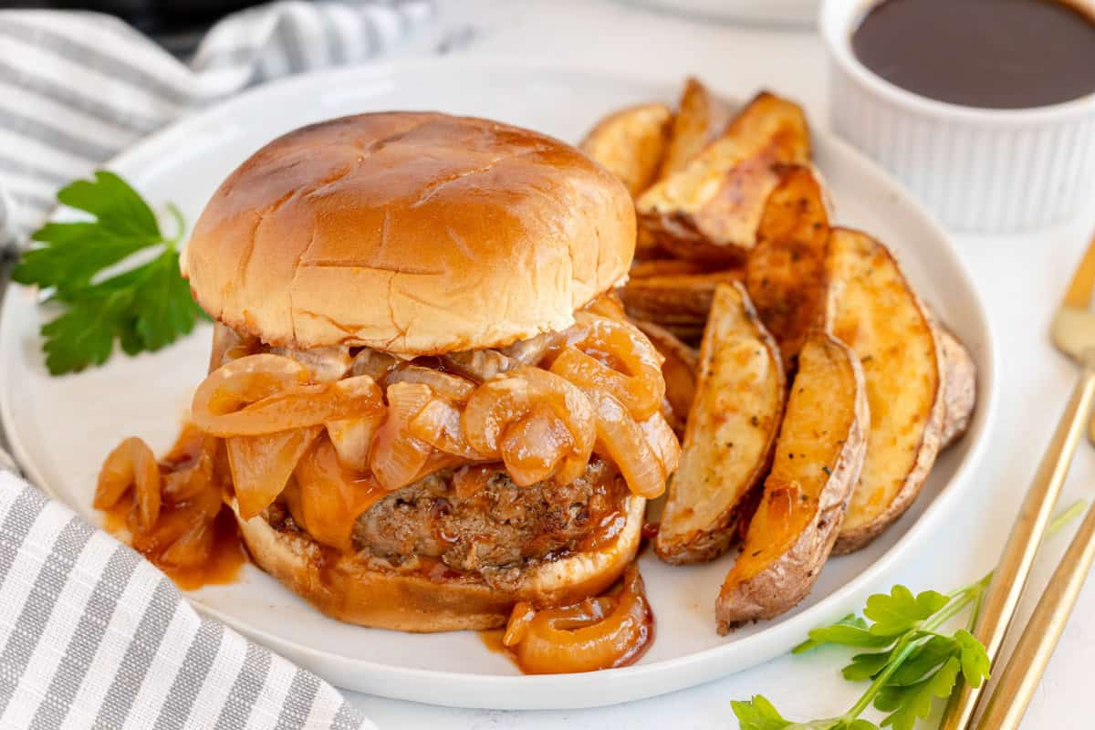 A burger topped with caramelized onions served with seasoned potato wedges on a white plate.