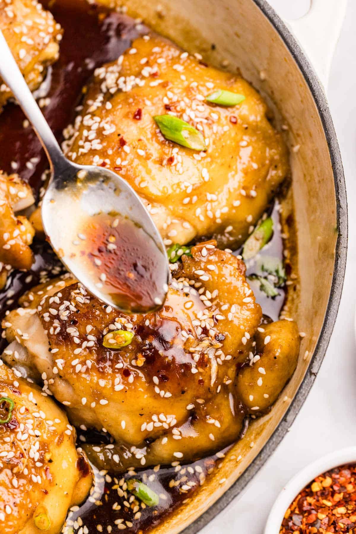 Spoon pouring glaze over chicken thighs coated in sauce, sesame seeds, and green onions inside a skillet.
