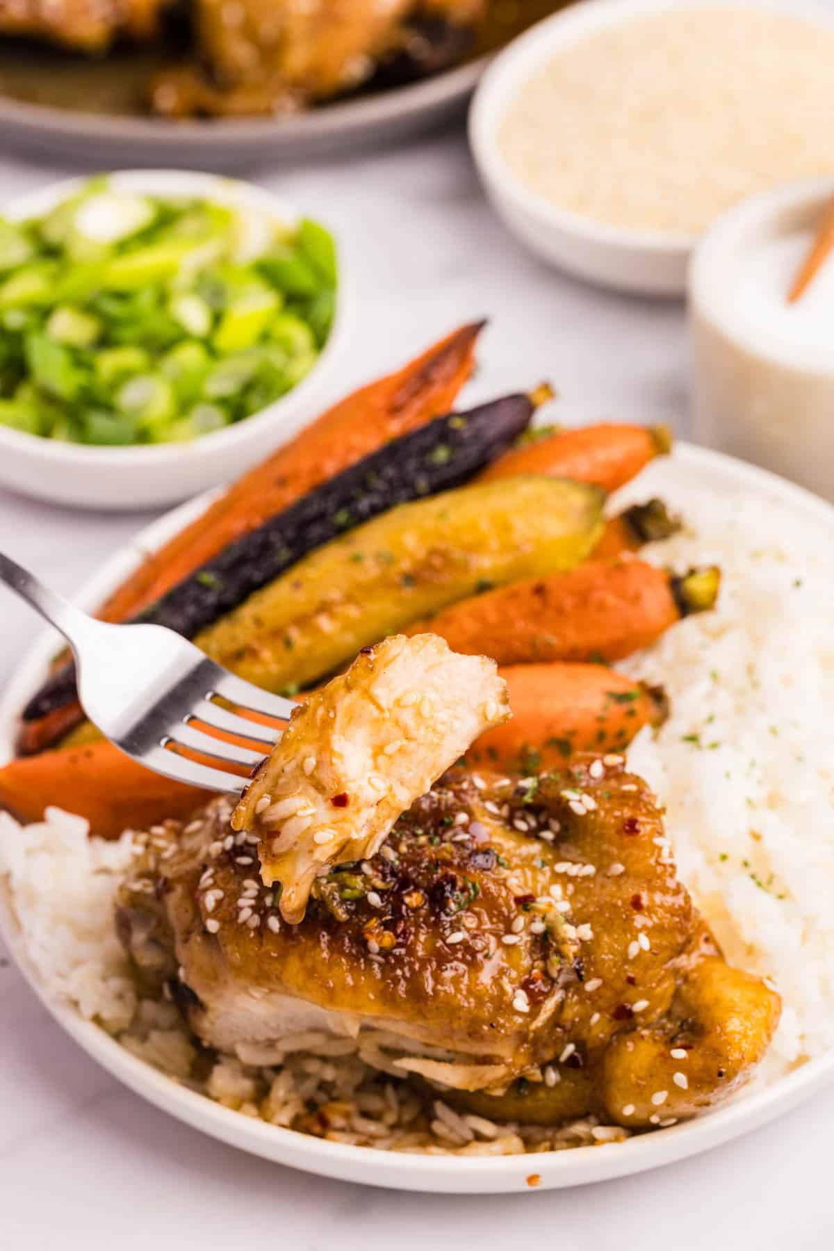 A fork holding a piece of glazed chicken over a plate of rice, roasted carrots, and more glazed chicken.