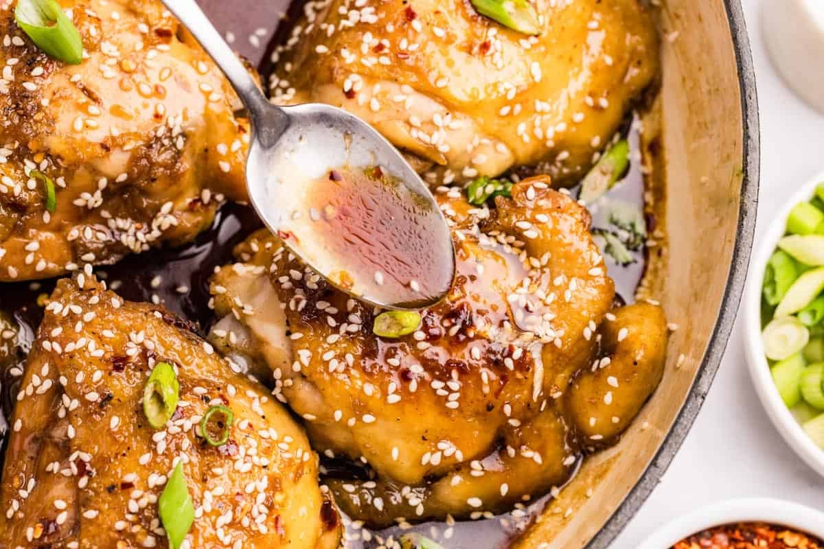 Chicken thighs in a skillet, sprinkled with sesame seeds and green onions, with sauce being spooned on top.