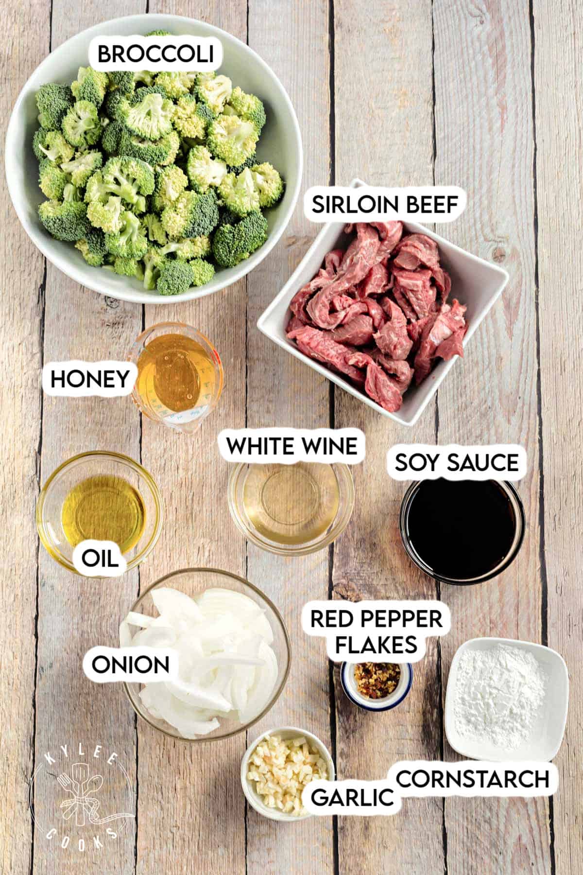 Overhead shot of ingredients for honey soy beef and broccoli, including bowls of broccoli, sliced sirloin beef, onions, garlic, soy sauce, honey, oil, white wine, cornstarch, and red pepper flakes, each labeled with text.