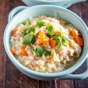 A bowl of chicken and rice soup with carrots and parsley garnish, served in a light blue ceramic bowl on a wooden surface.