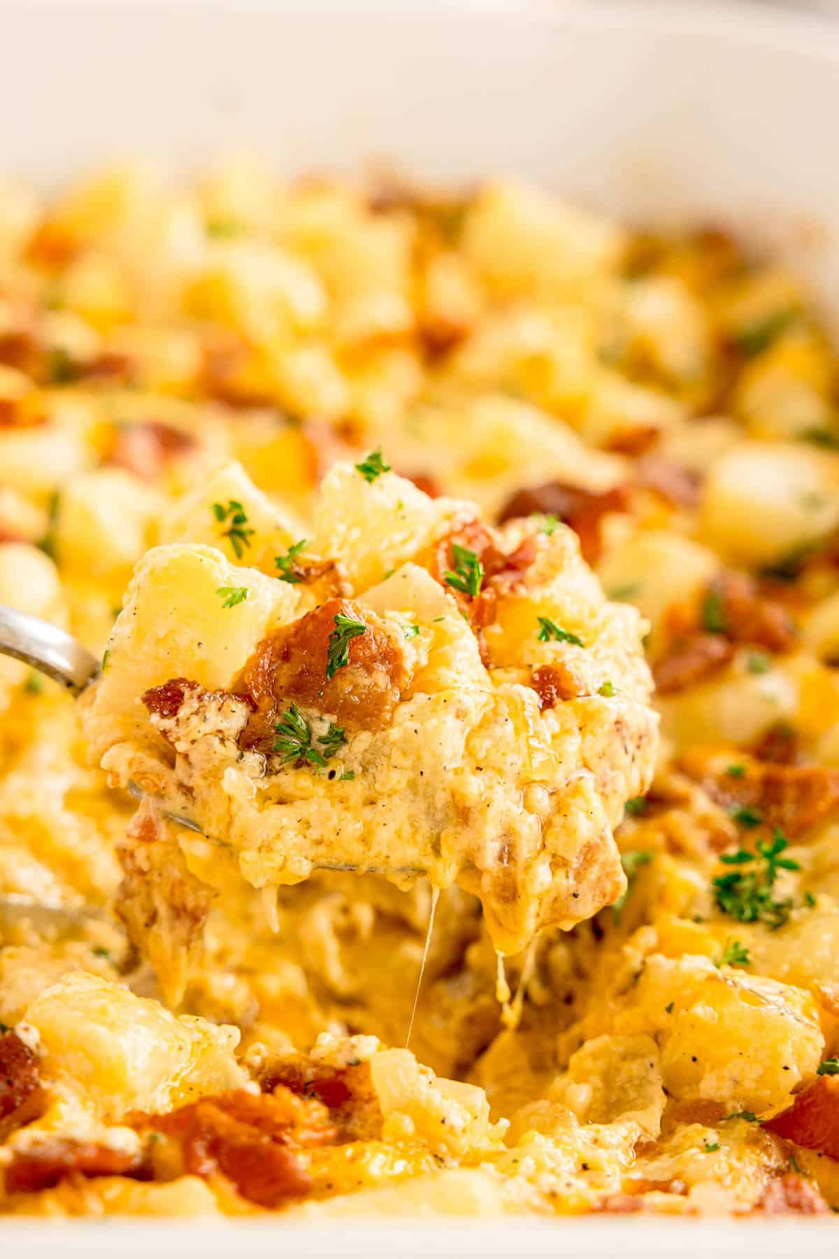 A spoonful of cheesy potato casserole with bacon being lifted from the dish, showing melted cheese stretching.