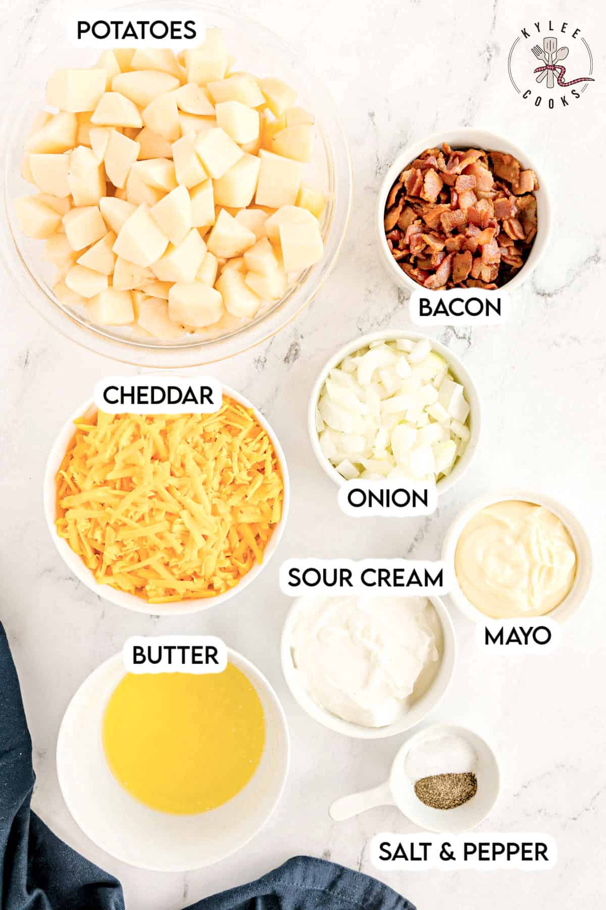 Bowls with chopped potatoes, shredded cheddar, bacon, onion, sour cream, mayo, melted butter, and a spoonful of salt and pepper. Text on the image says 'POTATOES', 'BACON', 'CHEDDAR', 'ONION', 'SOUR CREAM', 'MAYO', 'BUTTER', 'SALT & PEPPER'.