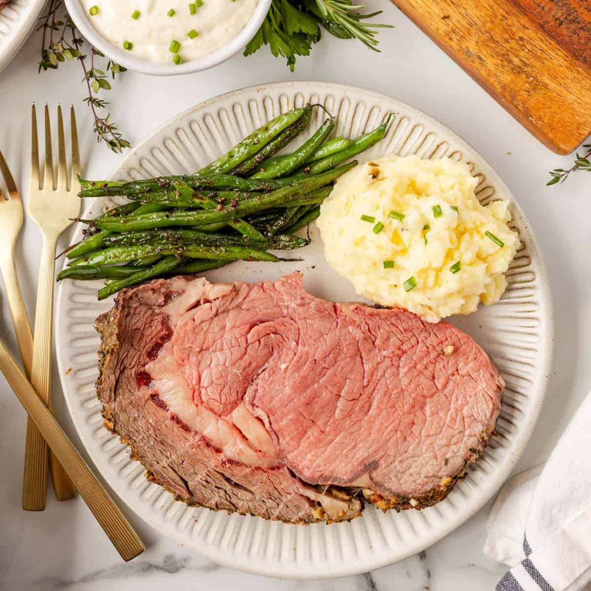 A plated meal with a slice of herb-crusted prime rib, mashed potatoes garnished with chives, and green beans.