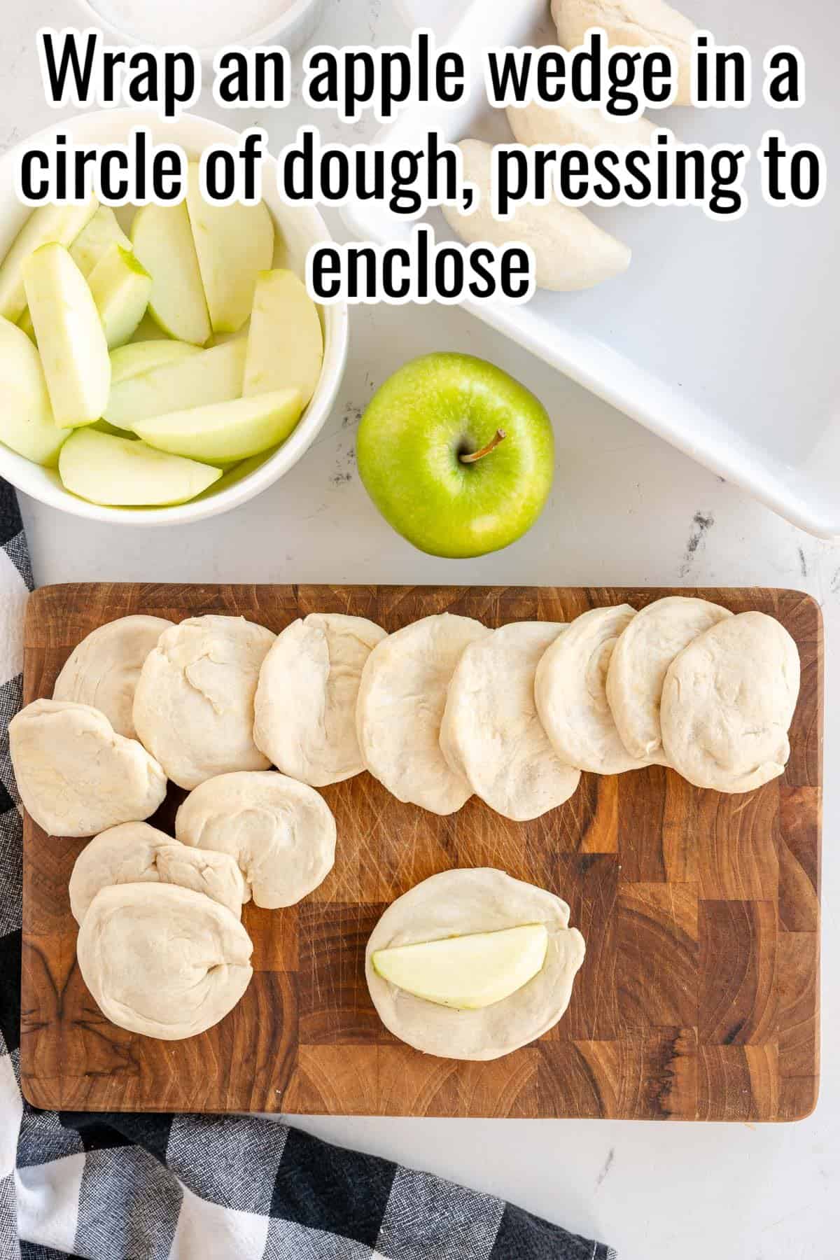 Apple wedges and dough are on a wooden cutting board, with one apple wedge partially wrapped. A green apple and a bowl with more apple wedges are beside the board. Text instructions are superimposed above.