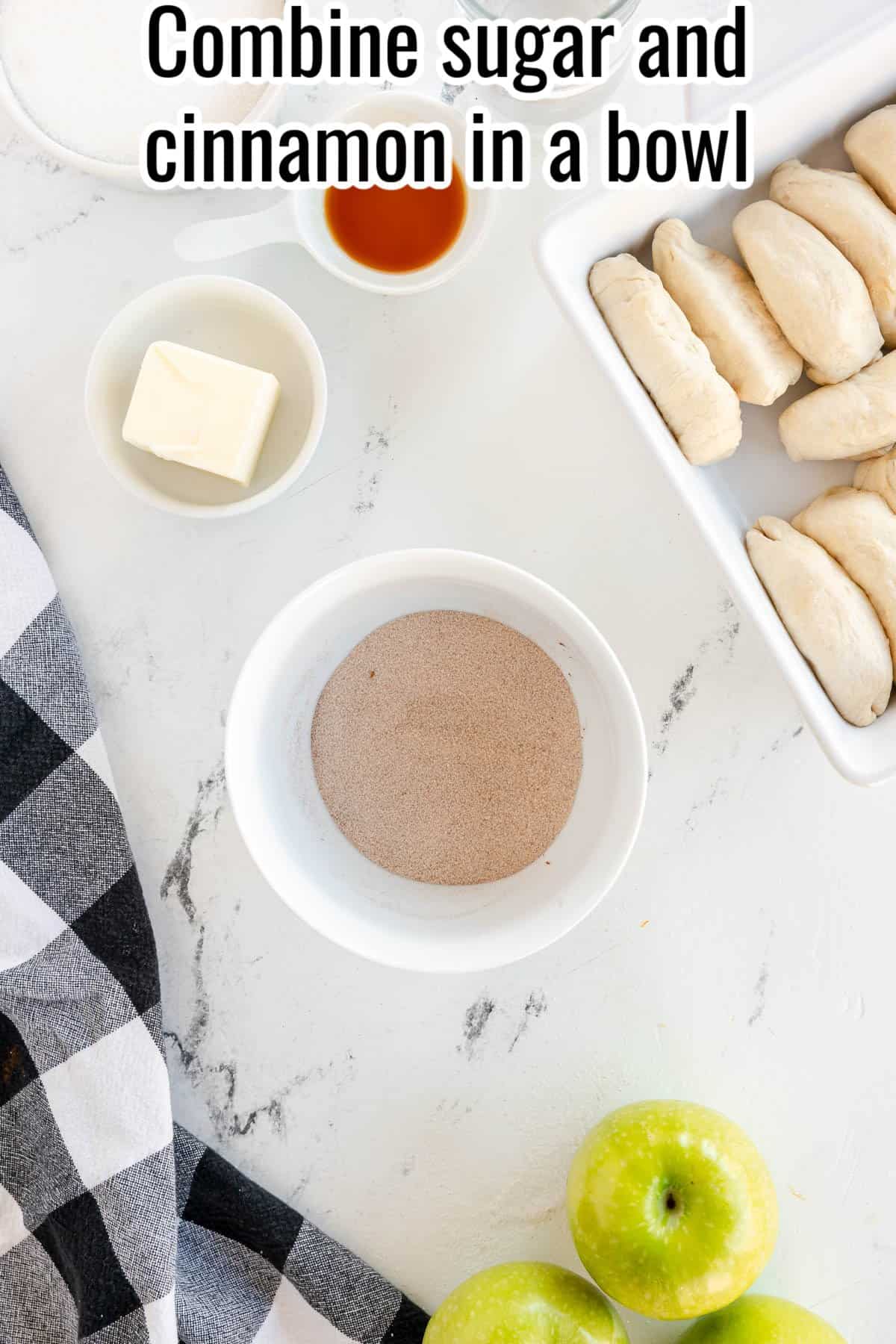 A bowl of sugar and cinnamon mixture, a small bowl of butter, a cup of vanilla extract, a baking dish with uncooked biscuits, a green apple, and a checkered cloth on a white countertop.