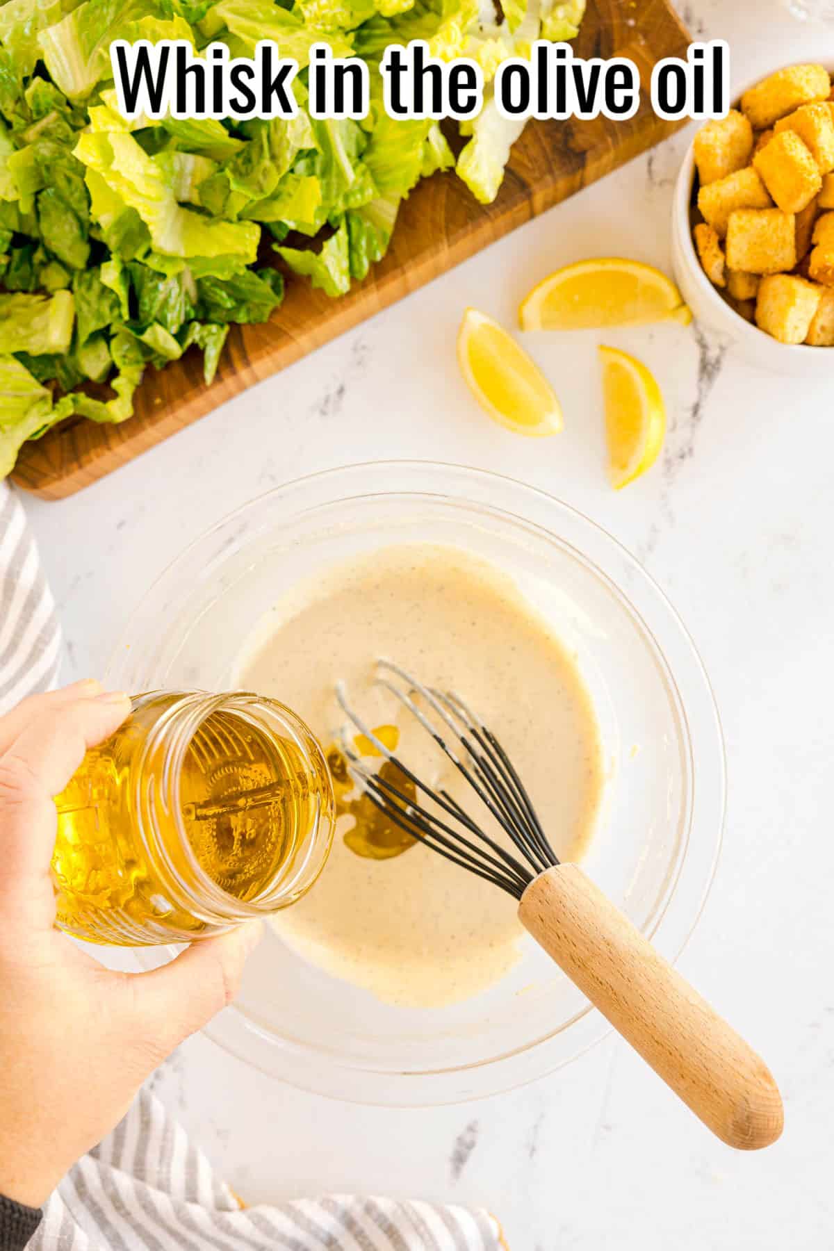Olive oil being poured into a bowl of dressing with a whisk inside. Text on the image says 'Whisk in the olive oil'.