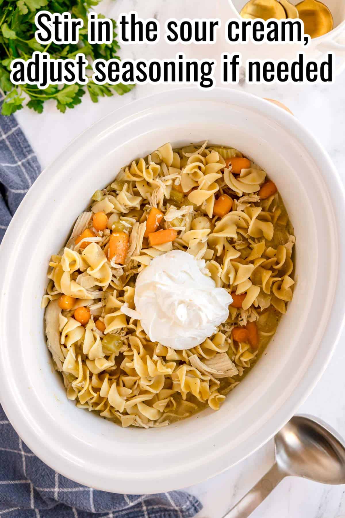 Slow cooker filled with chicken, noodles, and vegetables, with sour cream on top. Text on the image says 'Stir in the sour cream, adjust seasoning if needed'.