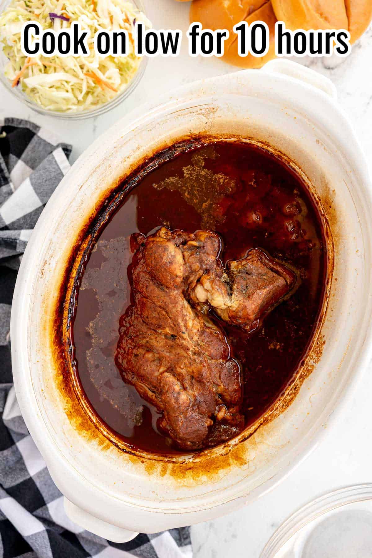 A cooked dish of pulled pork sits in a slow cooker with dark sauce, accompanied by a plaid towel, coleslaw, and bread rolls. Text at the top reads: "Cook on low for 10 hours.