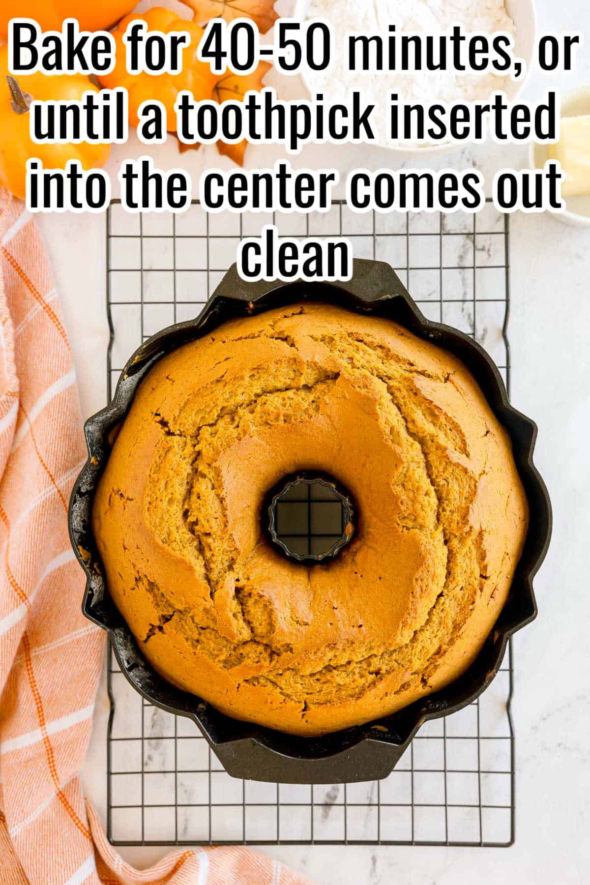 A baked pumpkin Bundt cake still in the pan on a cooling rack. Text on the image says 'Bake for 40-50 minutes, or until a toothpick inserted into the center comes out clean'.