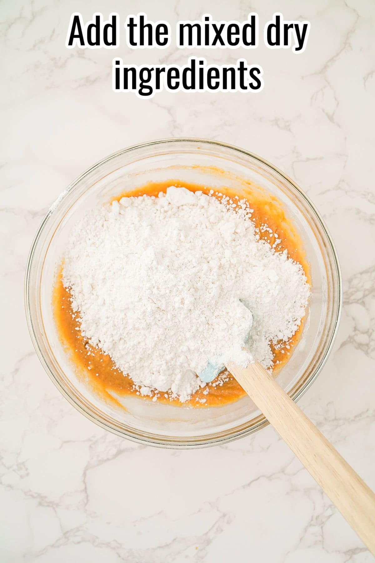 A mixing bowl with dry ingredients on top of pumpkin mixture. A spatula rests inside. Text on the image says 'Add the mixed dry ingredients'.