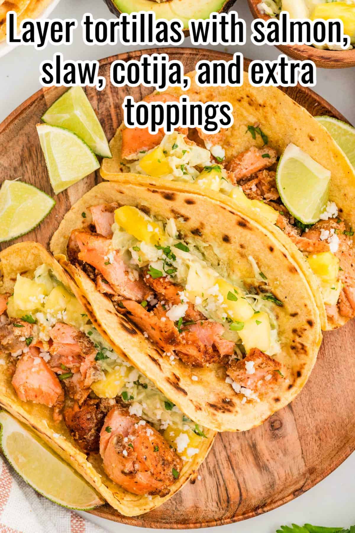 Tacos filled with salmon, slaw, pineapple, cotija cheese, and lime wedges. Text on the image says 'Layer tortillas with salmon, slaw, cotija, and extra toppings'.