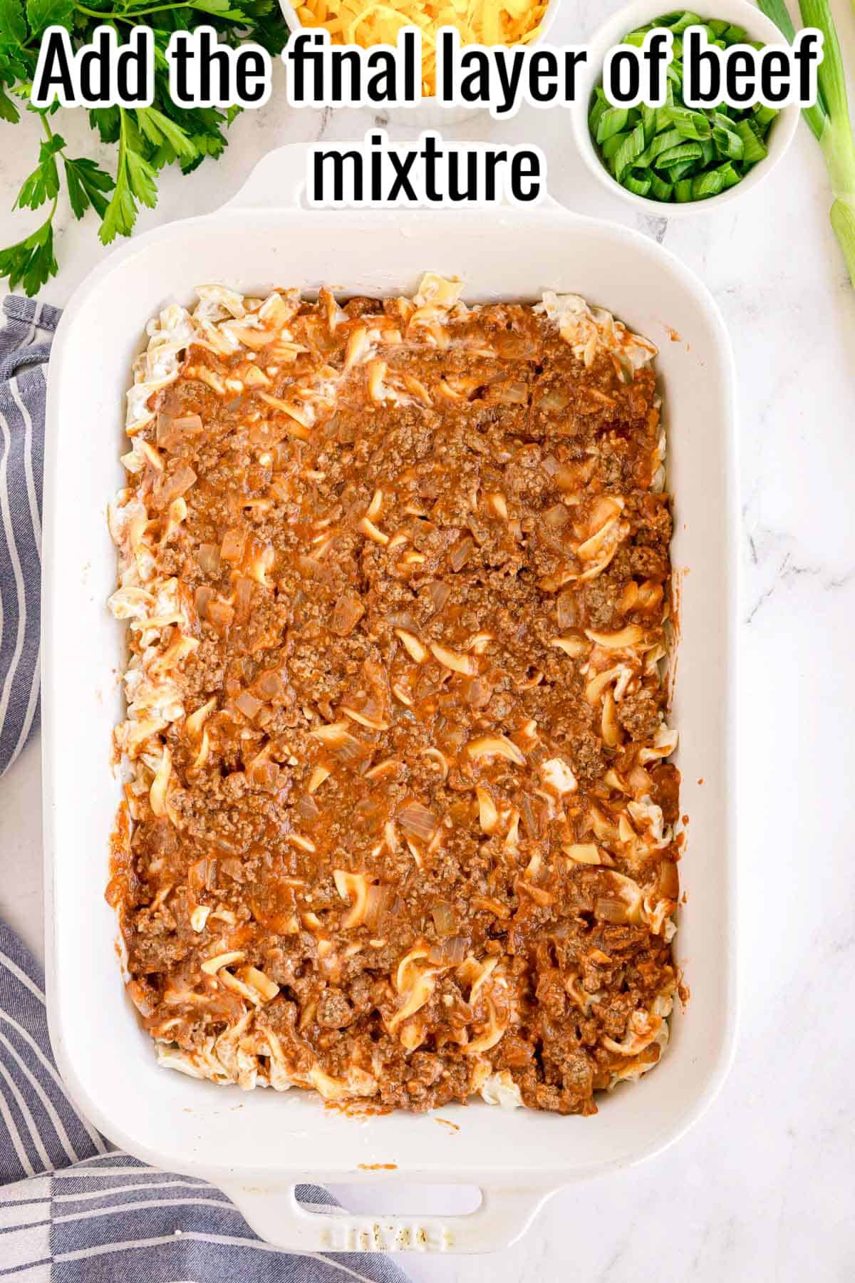 The final layer of beef mixture spread over the noodles. Text on the image says 'Add the final layer of beef mixture'