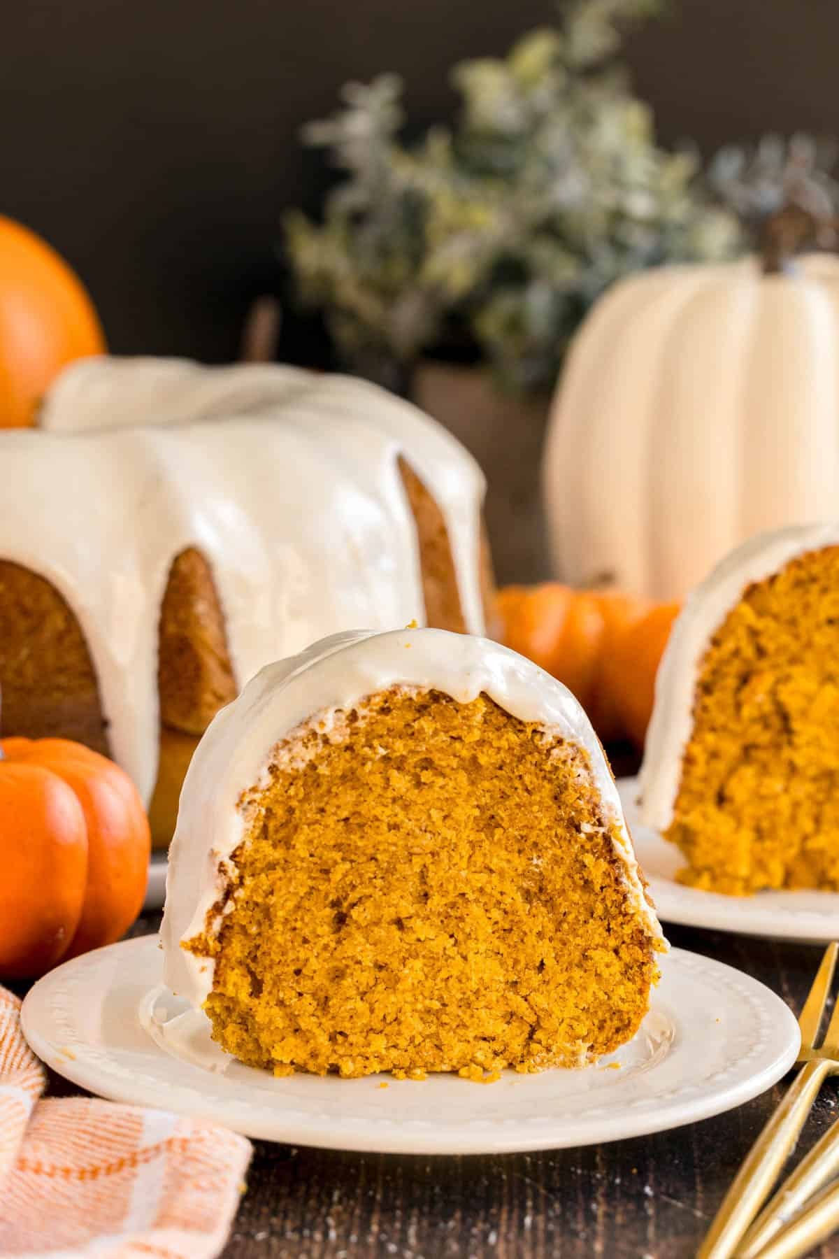 A slice of pumpkin bundt cake with cream cheese glaze on a plate, with the rest of the cake in the background.