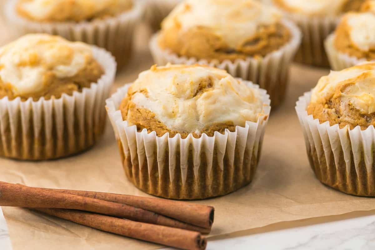 Pumpkin muffins with swirls of cream cheese in white paper liners, with cinnamon sticks on the side.