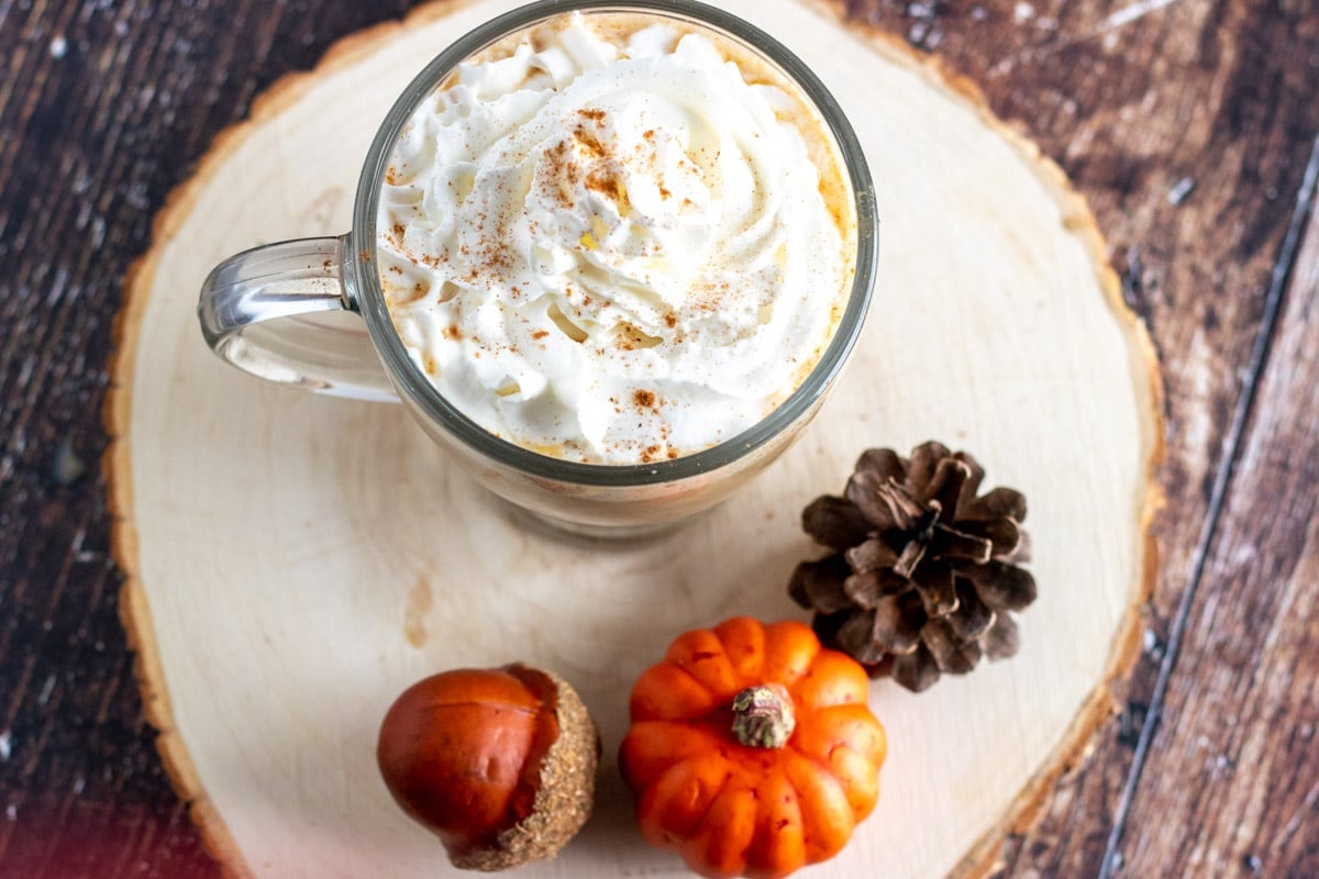 overhead shot of a cup of coffee topped with whipped cream, on a wood base with pumpkin decor