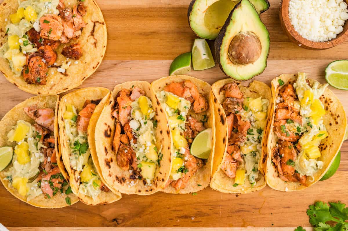 Six salmon tacos lined up on a wooden board, topped with pineapple slaw, cheese, and cilantro, with lime wedges and avocado nearby.