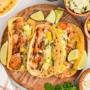 Three salmon tacos on a wooden plate, topped with pineapple, slaw, and cotija, with lime wedges on the side.