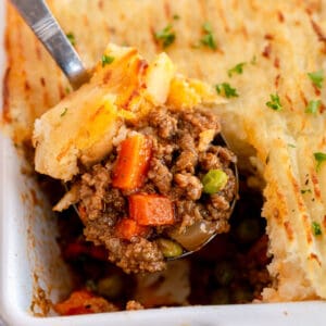 A serving spoon lifts a portion of shepherd's pie with ground meat, carrots, peas, and mashed potato topping from a baking dish.