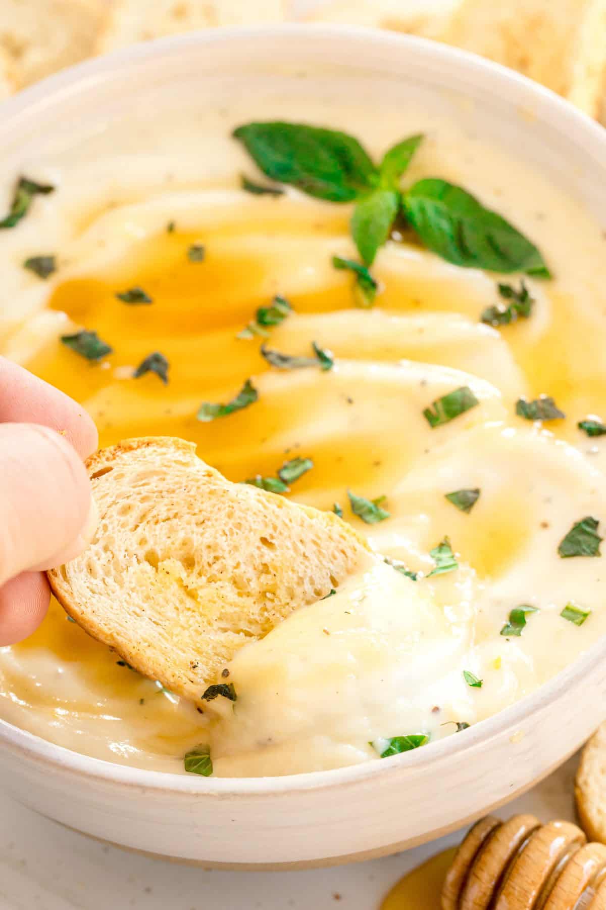 A hand dips a slice of bread into a bowl of creamy dip topped with herbs and a drizzle of honey.